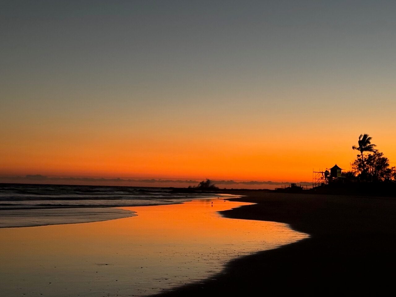 Sunset beach scene with orange sky and wet sand reflecting the colors; dark silhouettes of a lifeguard tower and palm trees on the right.