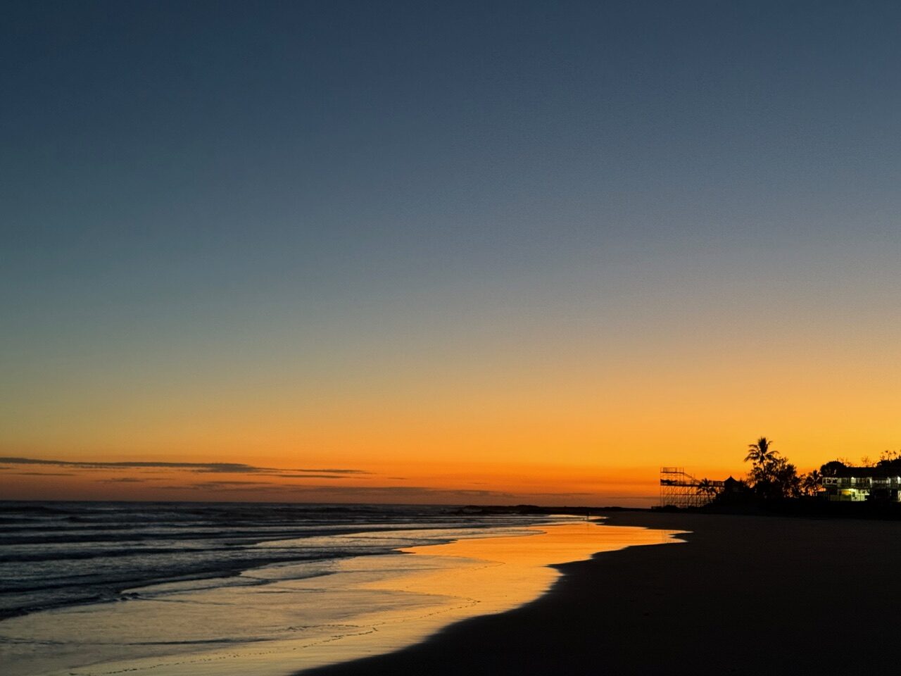 Sunset over a quiet beach with an orange horizon, reflecting on wet sand, silhouettes of palm trees and buildings on the right.