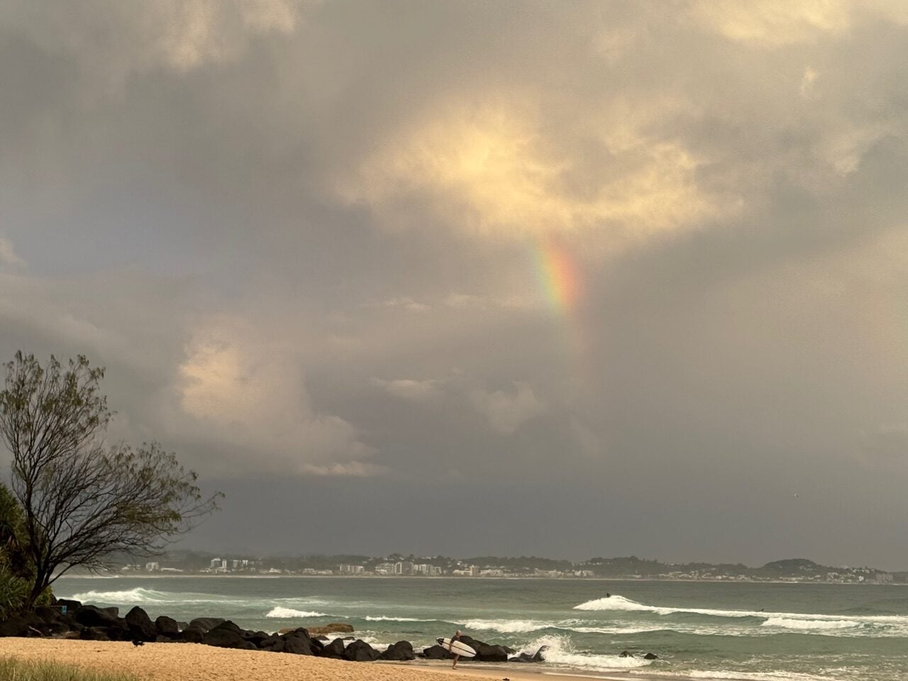 Rainbow arcing over a choppy sea and sandy beach with rocks and a tree on the left side of the frame, during a cloudy sky near sunset.