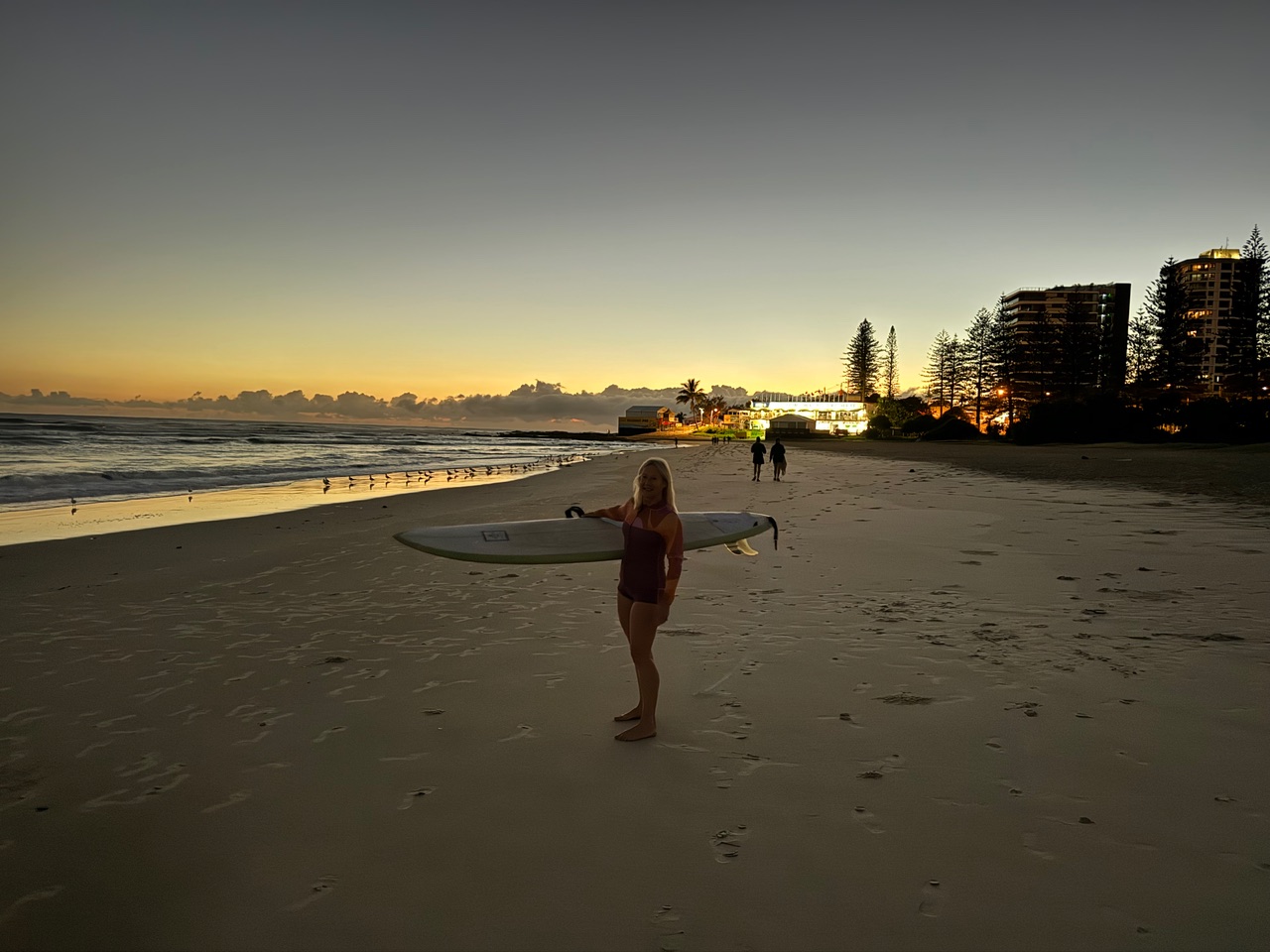 Woman on a sandy beach at sunset, holding a surfboard under her arm, with waves and buildings in the distance.