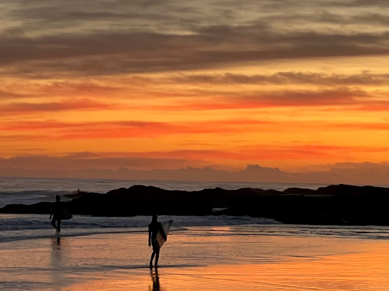 Two surfers walking along the wet sand at sunset with an orange sky and dark rocks in the distance.