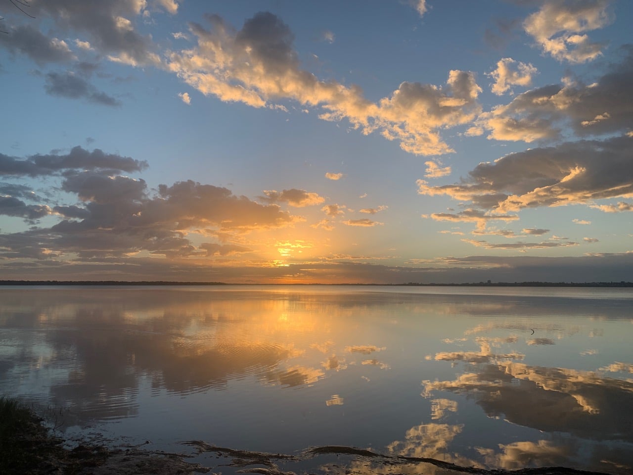 Sunset over a calm lake with a golden sky and fluffy clouds reflected in the still water, shoreline in the distance.