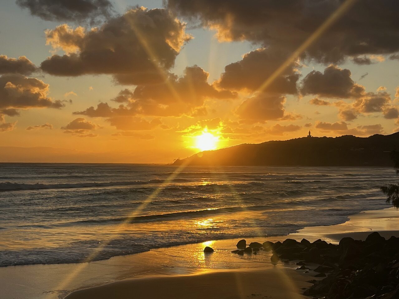 Sunset over the ocean with golden reflections on wet sand; rocky shore and distant headland in silhouette on the horizon.