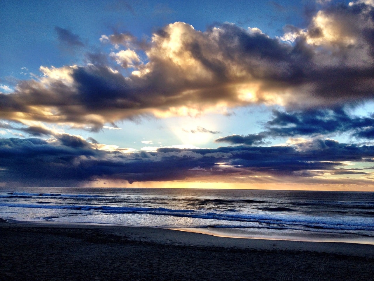 Sunset over the ocean with dark clouds and a warm golden glow near the horizon, gentle waves washing onto a sandy beach.