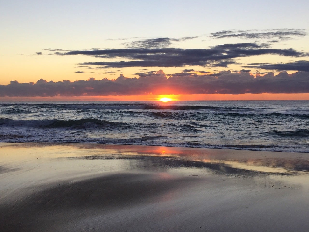 Sunset at the beach with an orange glow on the horizon, calm waves, and the wet sand reflecting the sky’s colors.