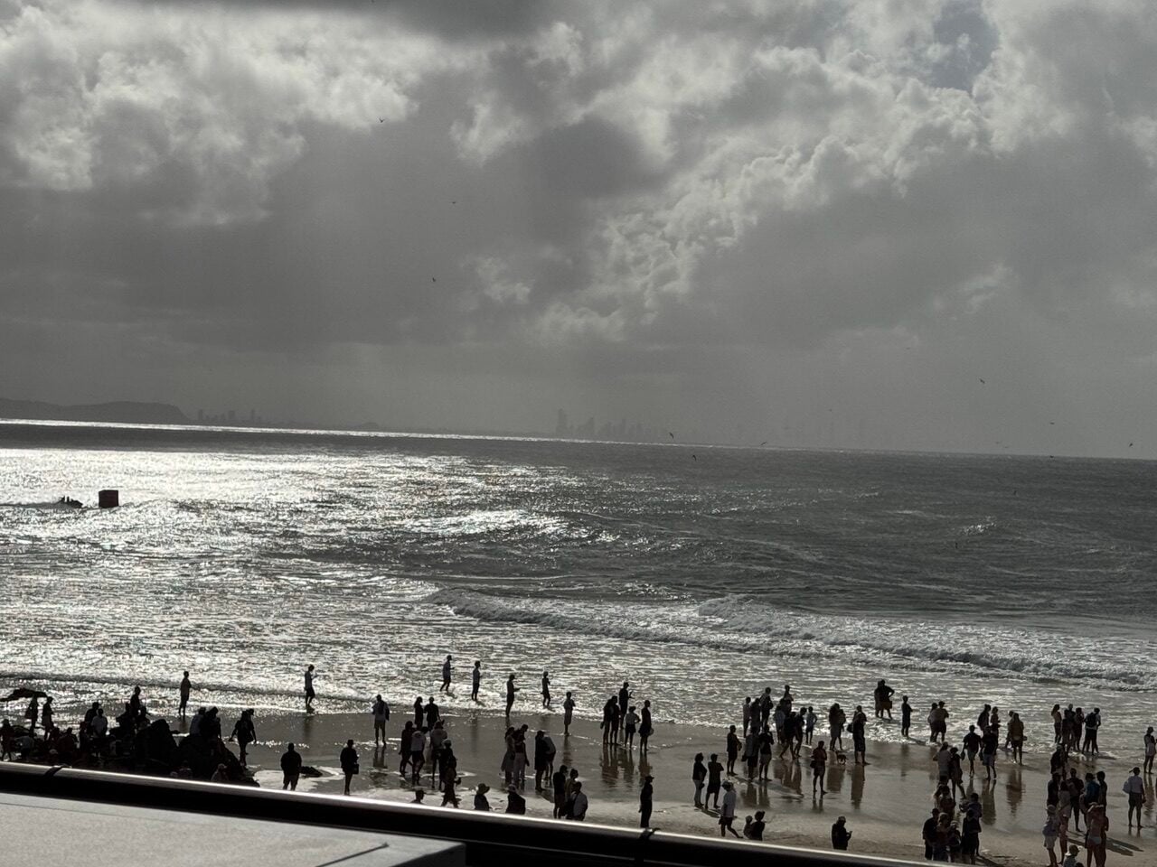 Crowded beach with many people along the shoreline, waves lapping in, and a distant city skyline under a cloudy sky.
