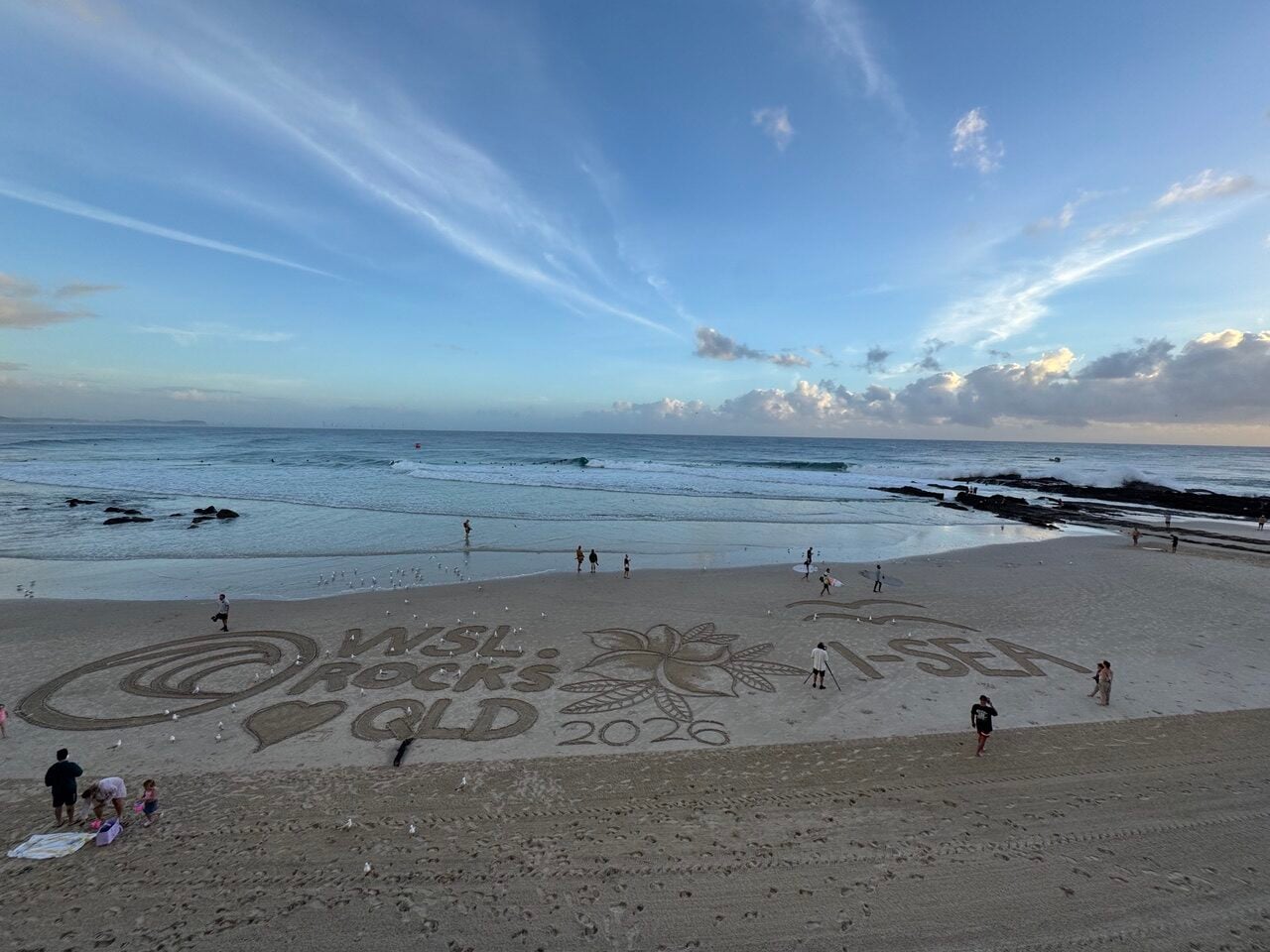 Sunny beach with a large sand drawing featuring a palm tree and the year 2026, with people walking along the shore and seagulls nearby.