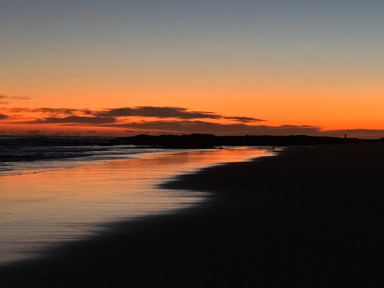 Sunset over a calm beach with orange sky and reflection on the water, dark silhouette of rocks and shore.