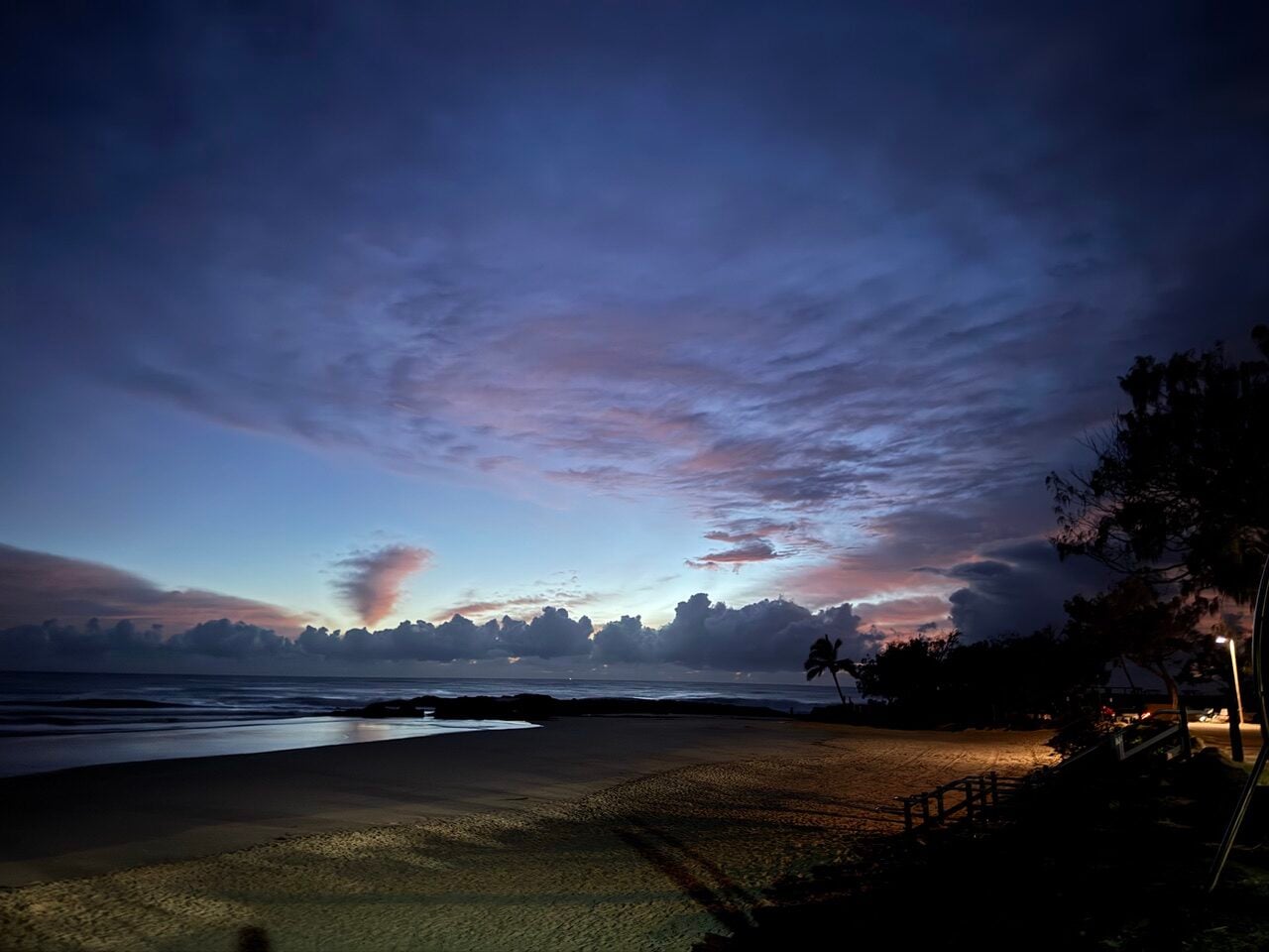 Dusk on a tropical beach with calm water, soft clouds, and palm trees along a lit path on the right.