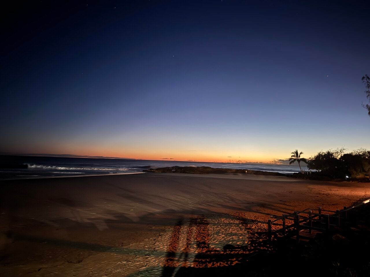 Twilight beach scene with a calm ocean, orange horizon, and palm trees, silhouettes on the sand and a wooden railing on the right edge.