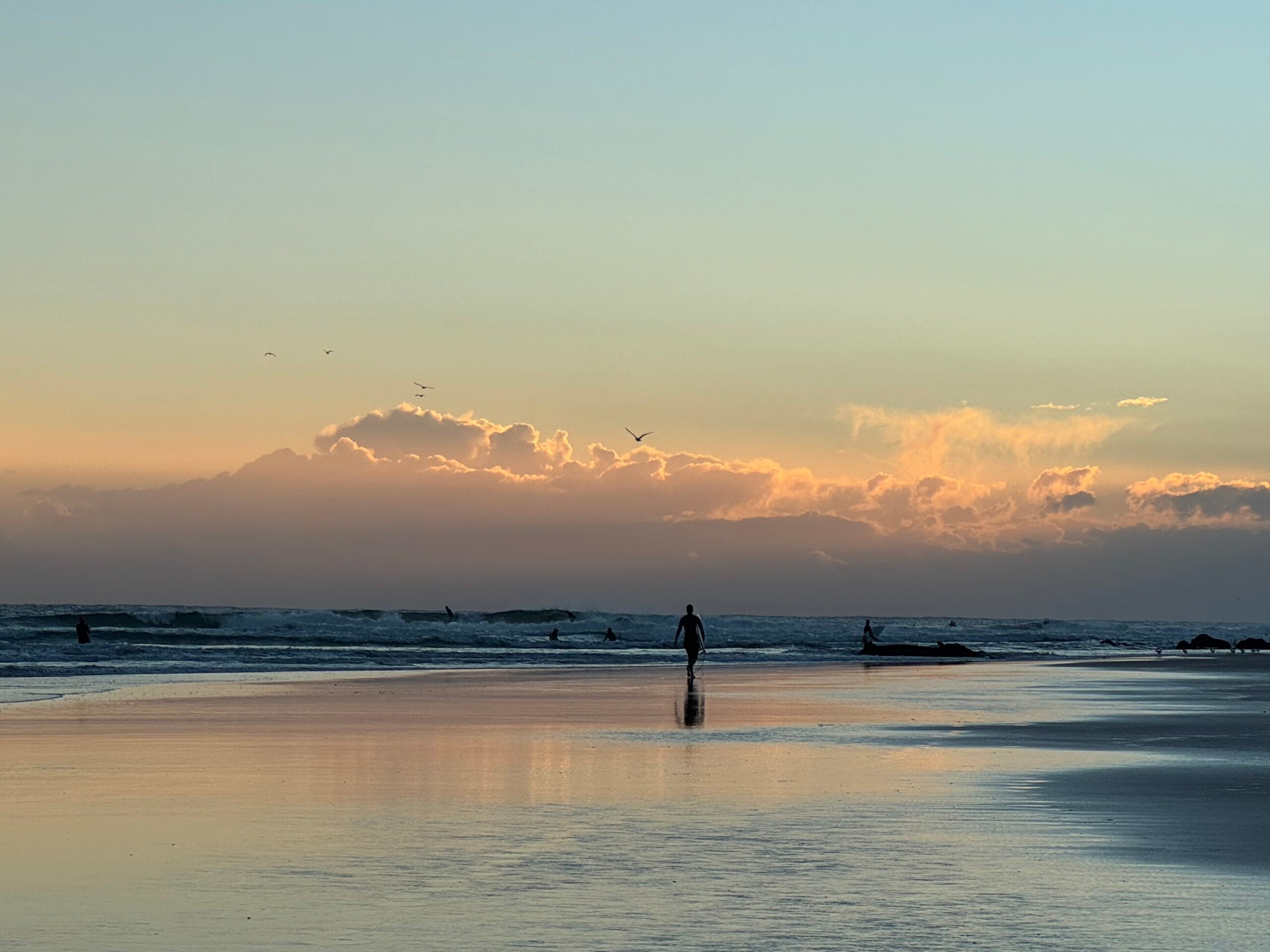 Beach at sunset with a pastel sky, gentle waves, and a few people wading in the shallow water on the shore.