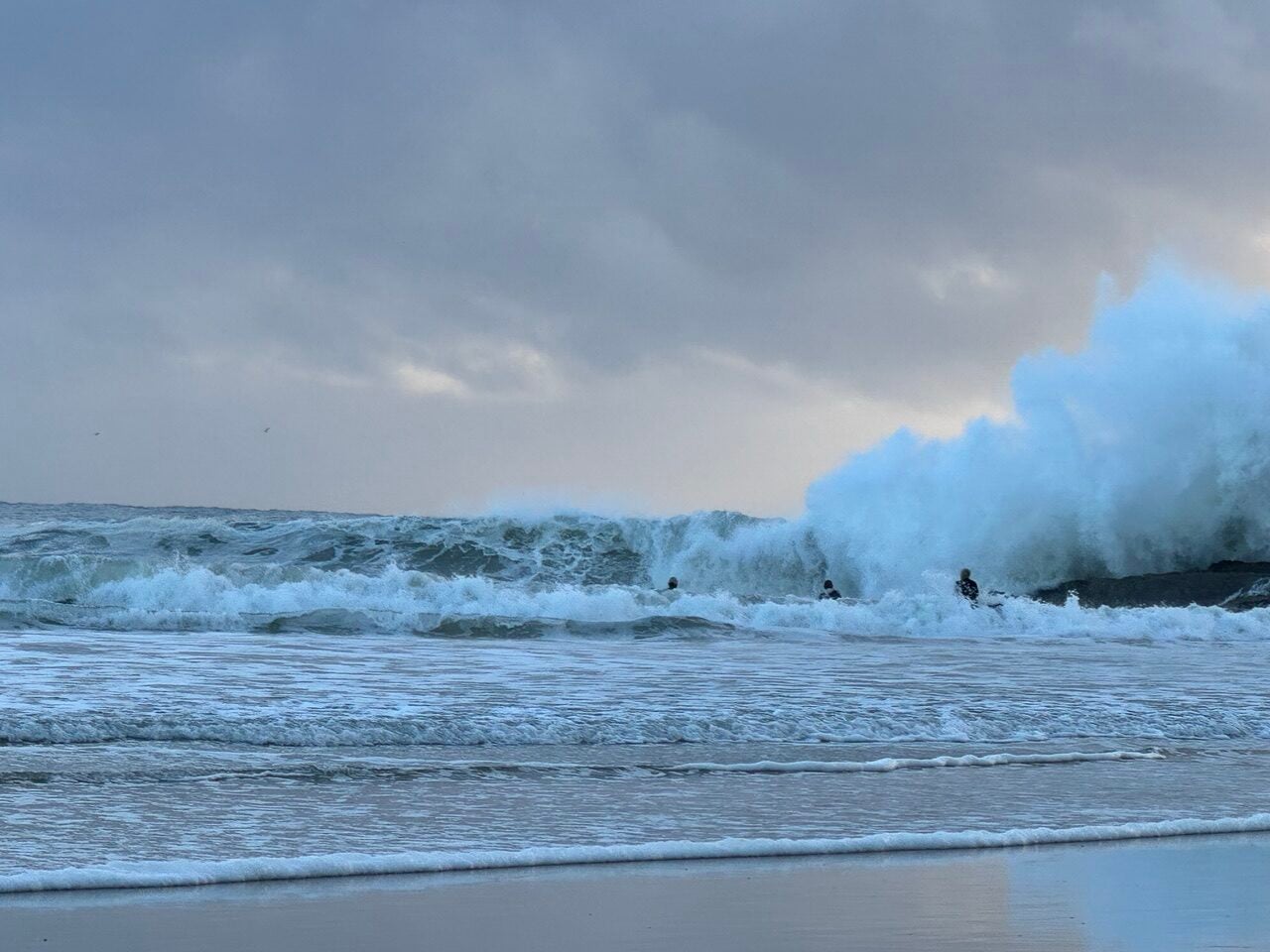 Large ocean wave breaking near a sandy shore with surfers visible in the water beneath a cloudy sky, beach in the foreground.