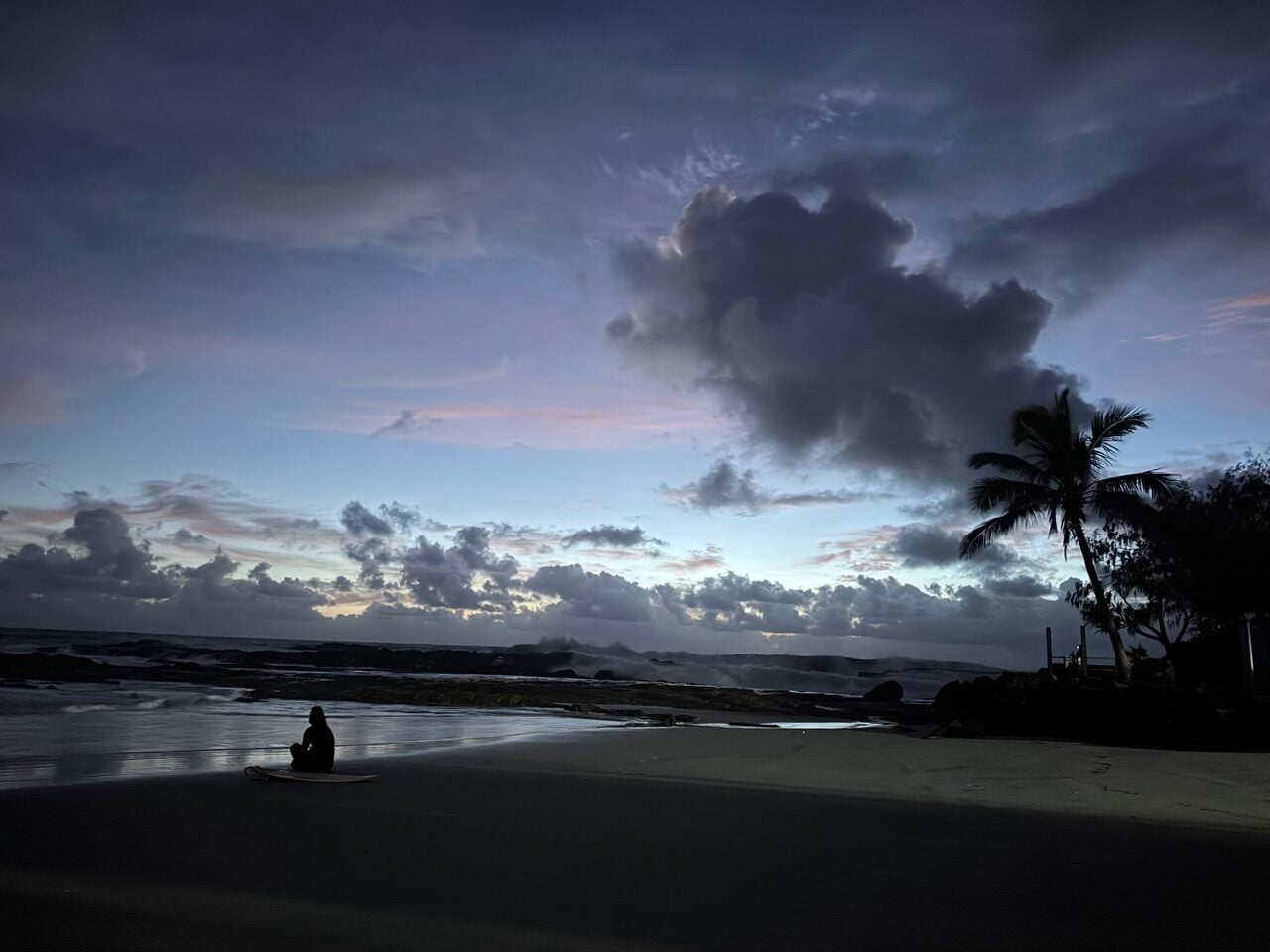 Silhouette of a person sitting on a surfboard on a wet beach at twilight, with a palm tree on the right and a cloudy sky.