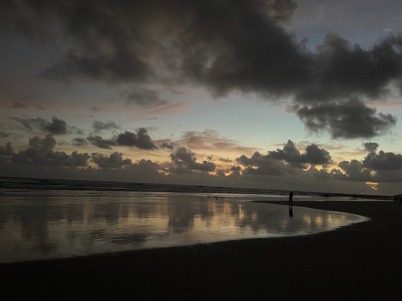 Silhouette of a person walking along a reflective beach at sunset with dark clouds overhead.