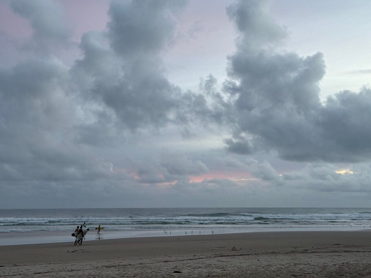 Overcast beach at dusk with a few surfers walking along the wet sand toward the waves, carrying their boards.