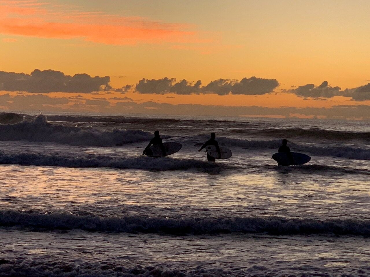 Three silhouetted surfers with their boards enter the ocean at sunset, orange sky reflecting on rolling waves.