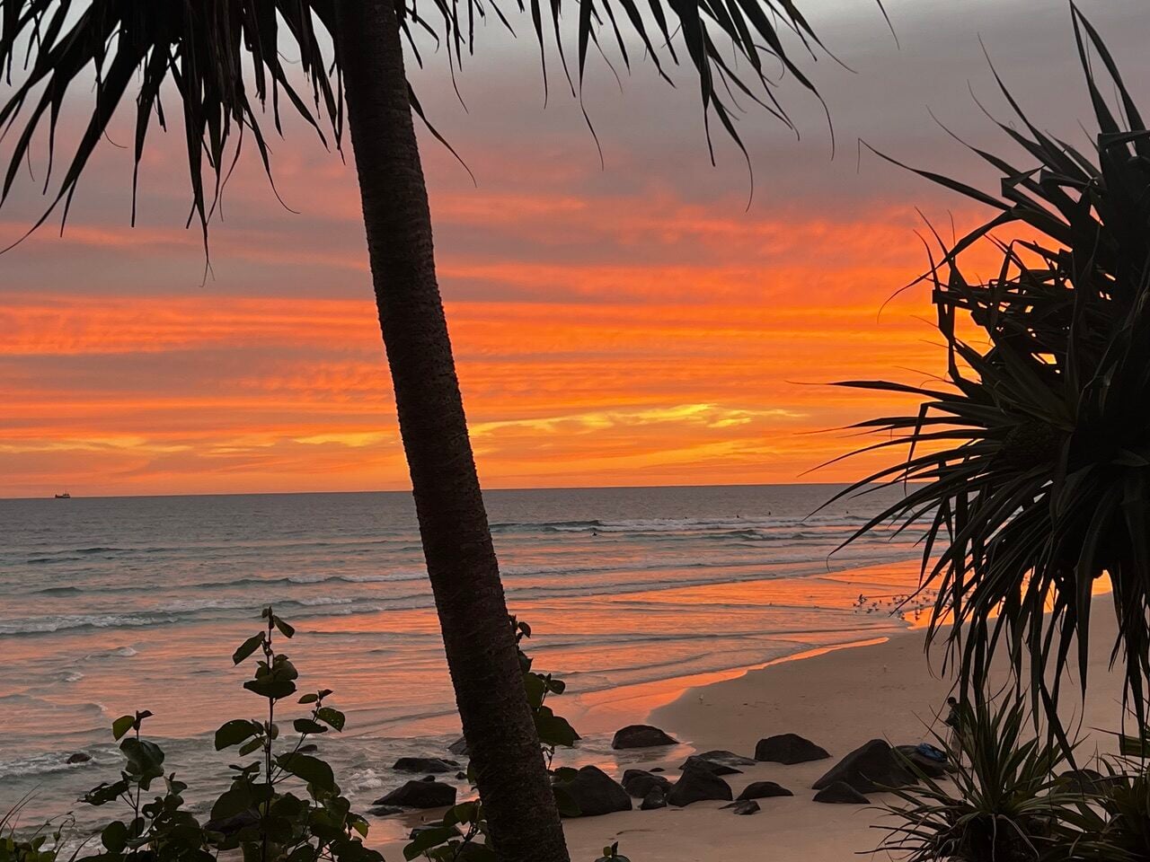 Sunset over the ocean at a tropical beach, silhouettes of palm trees and rocks along the shoreline.
