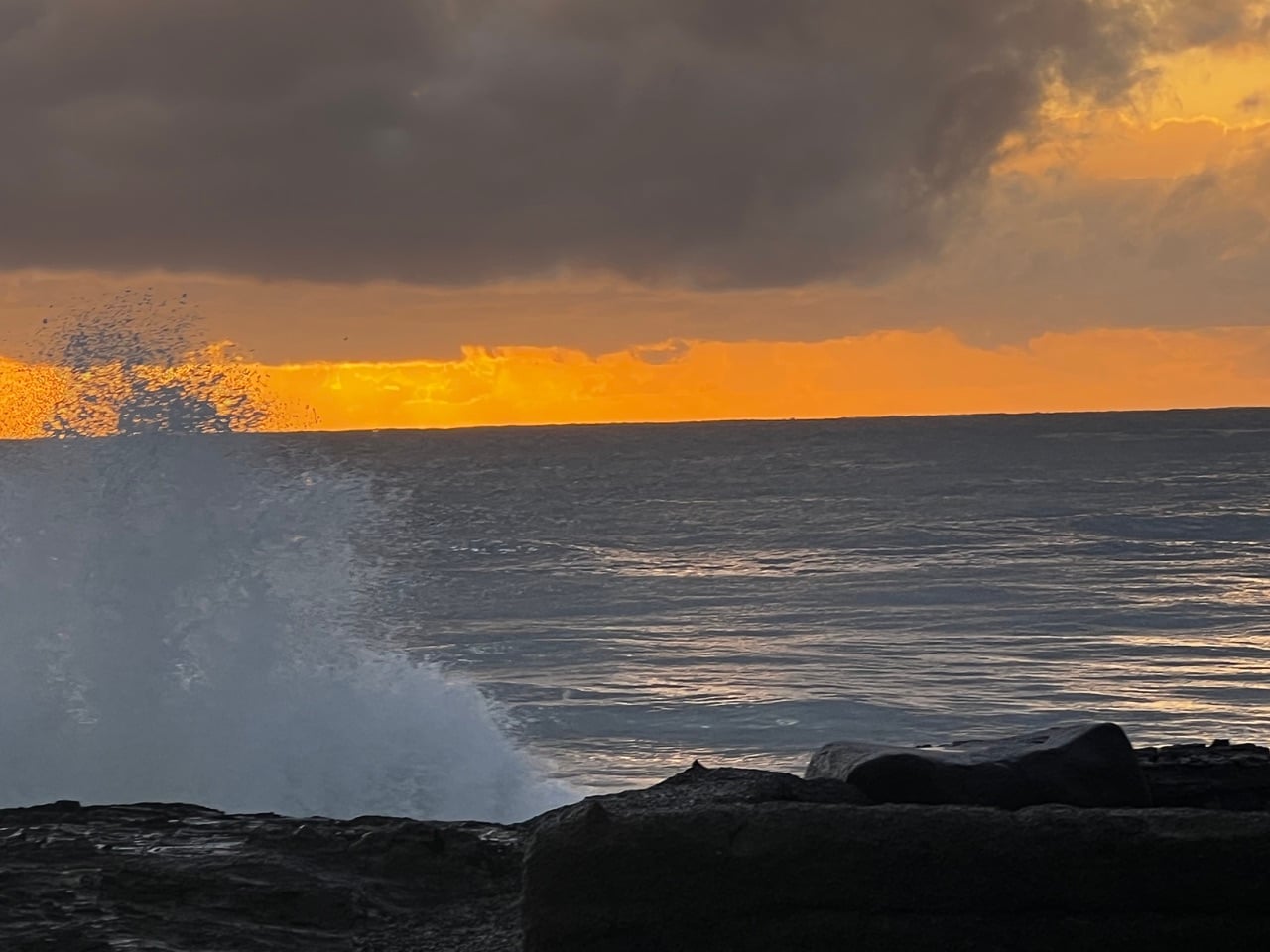 Crashing wave against rocky shore at sunset, orange sky reflecting on calm sea.