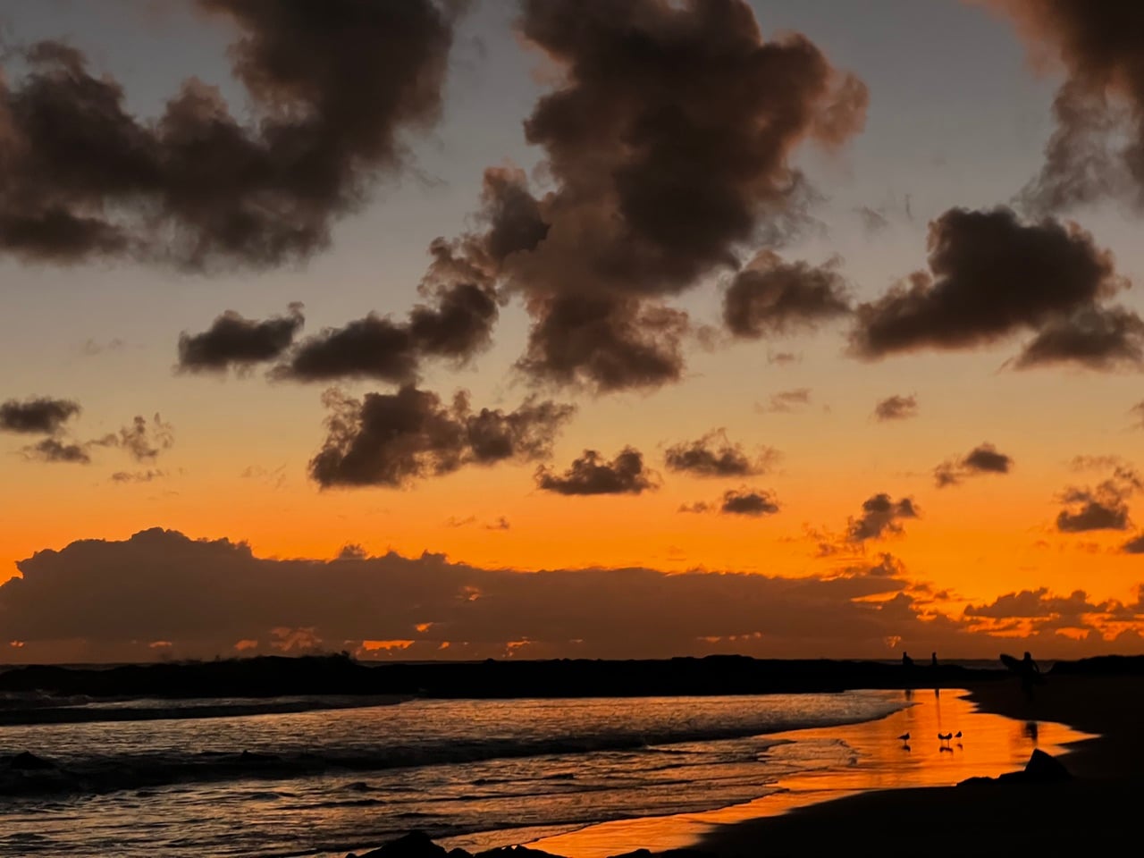Sunset over a calm sea with an orange sky and dark clouds; silhouettes of people on the shore.