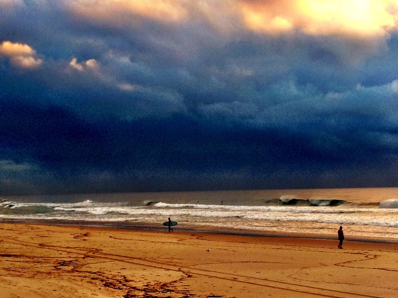 Stormy sky above a empty beach; a surfer with a board stands near the water while a person walks along the shore.