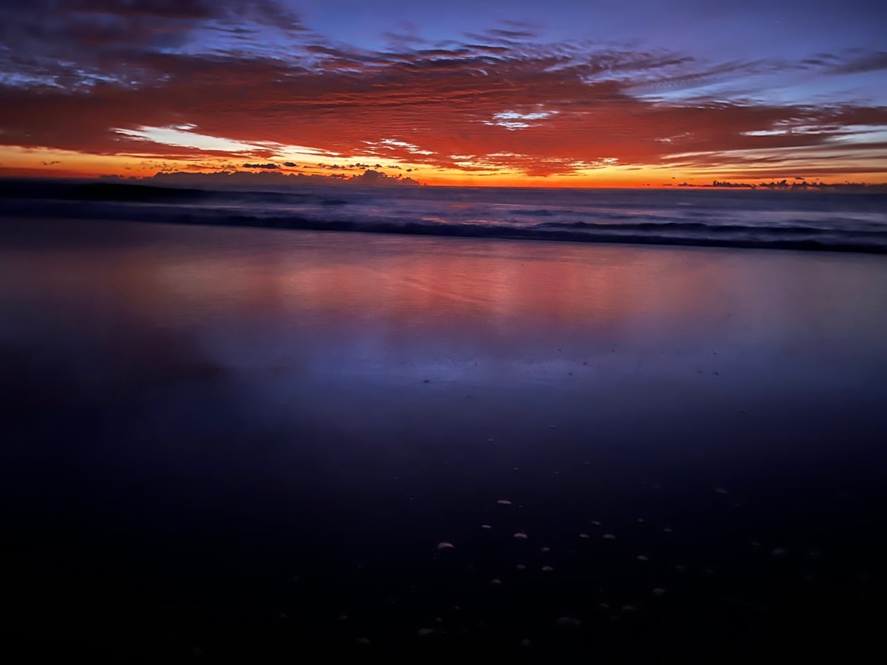 Sunset over calm ocean with orange horizon and purple-blue sky reflected on wet sand or water surface in the foreground.