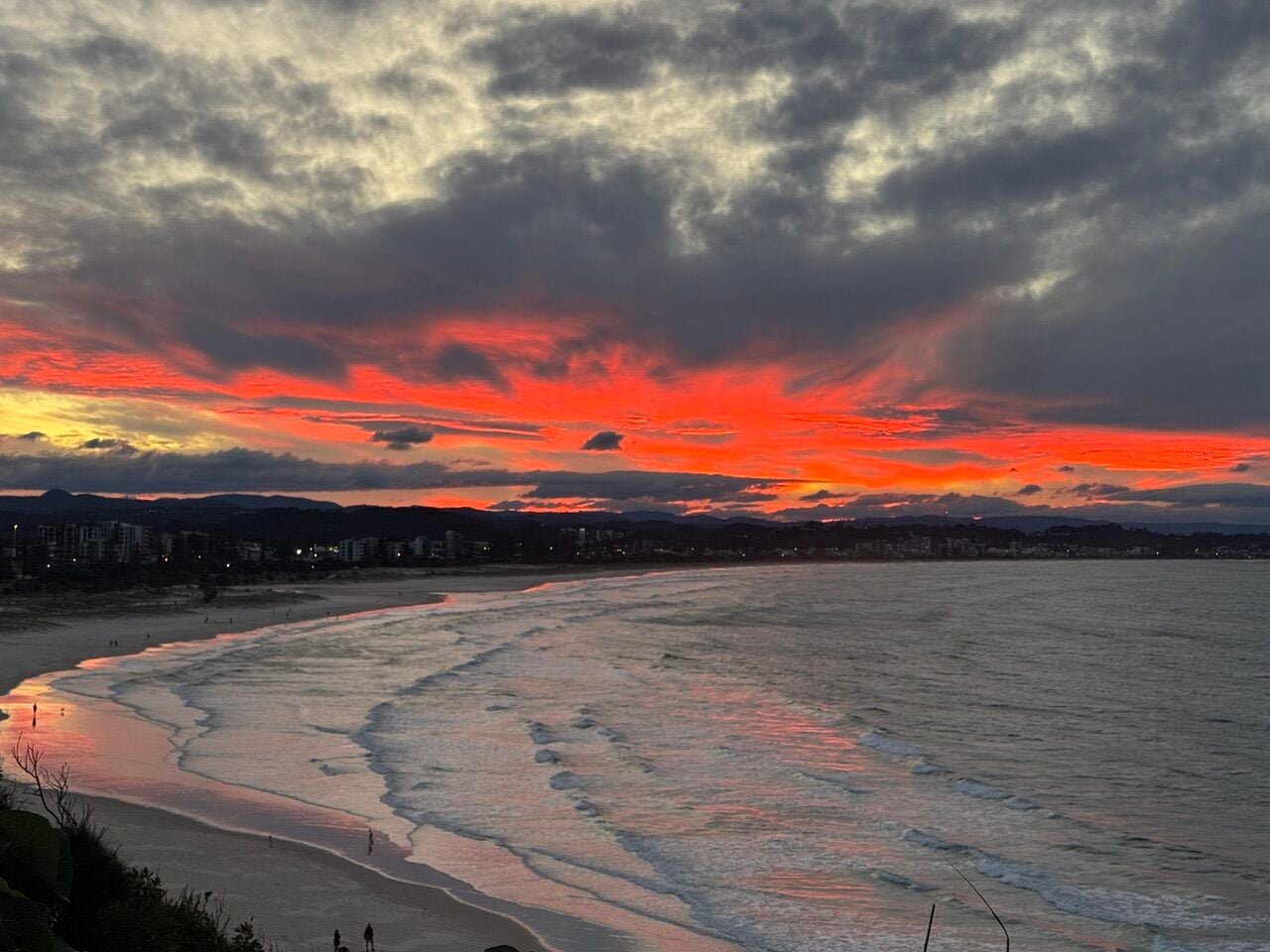 Sunset over a beach with bright red clouds reflecting on the wet sand and calm ocean, a distant shoreline with buildings under a dark cloud-filled sky