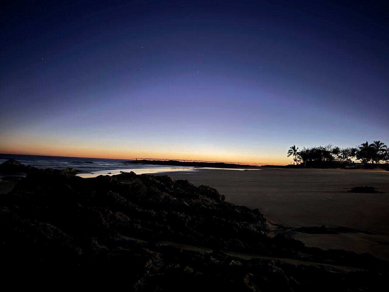 Twilight beach scene with dark rocks in the foreground, calm sea, orange glow on the horizon, and silhouetted palm trees on the right.