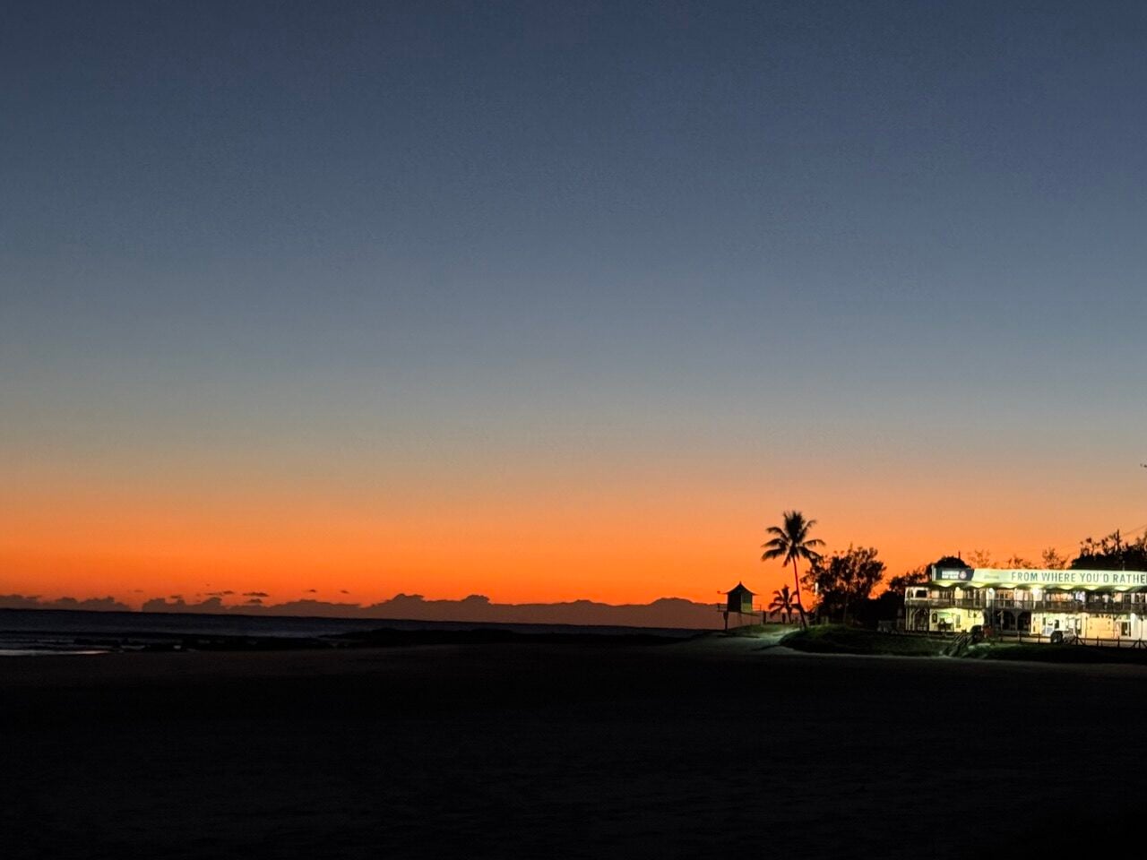 Sunset over a quiet beach with an orange horizon; a lit seaside restaurant and palm trees sit on the right.