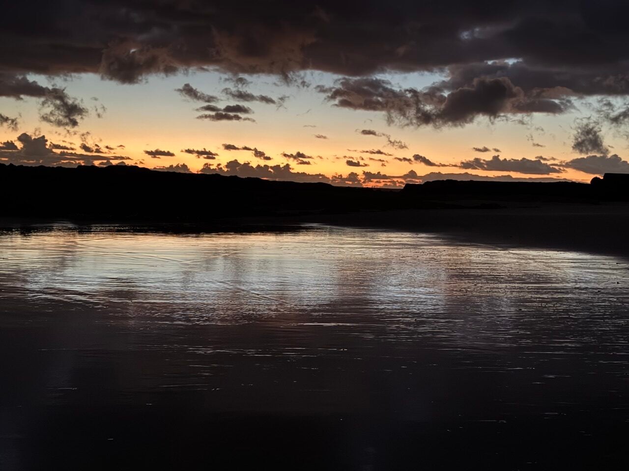 Sunset over calm coastal water with a dark silhouette of land and scattered clouds on the horizon.