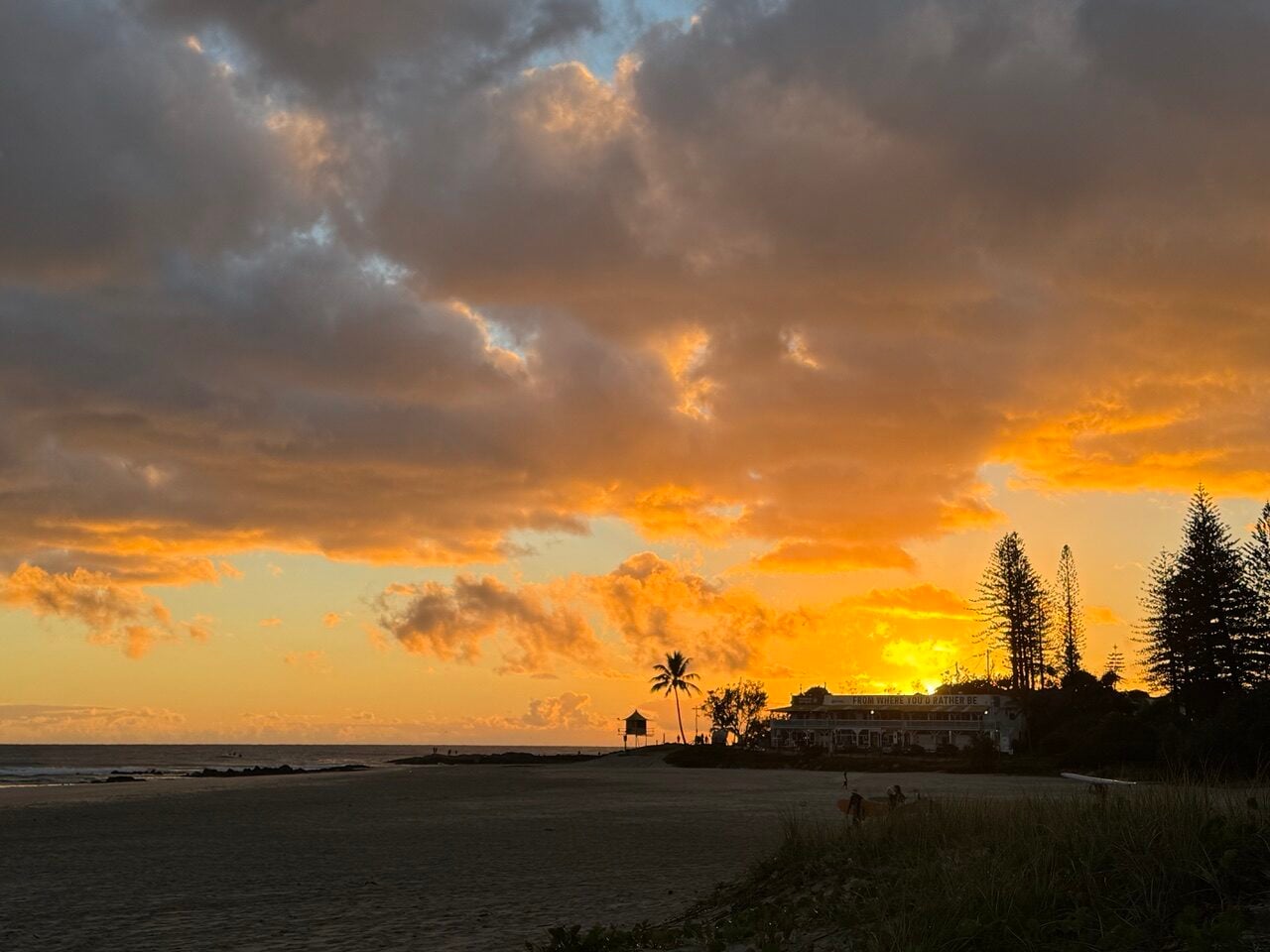 Sunset over a calm beach with orange sky, palm tree silhouettes, and a quiet building near the shoreline.
