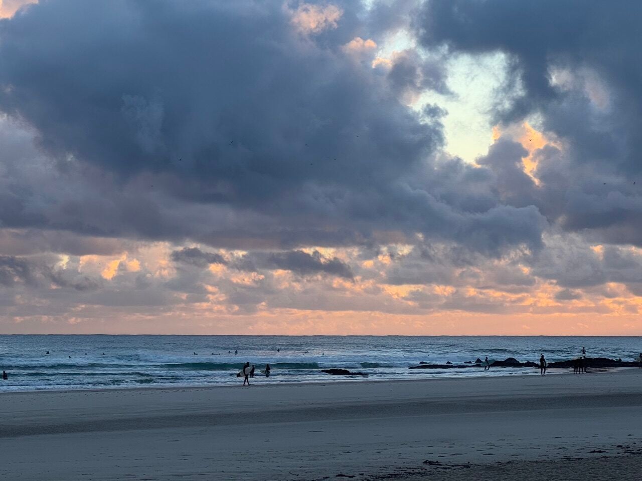Beach at dusk with heavy clouds and a pink-orange horizon; people walk along the wet sand by the water and rocks on the shore.