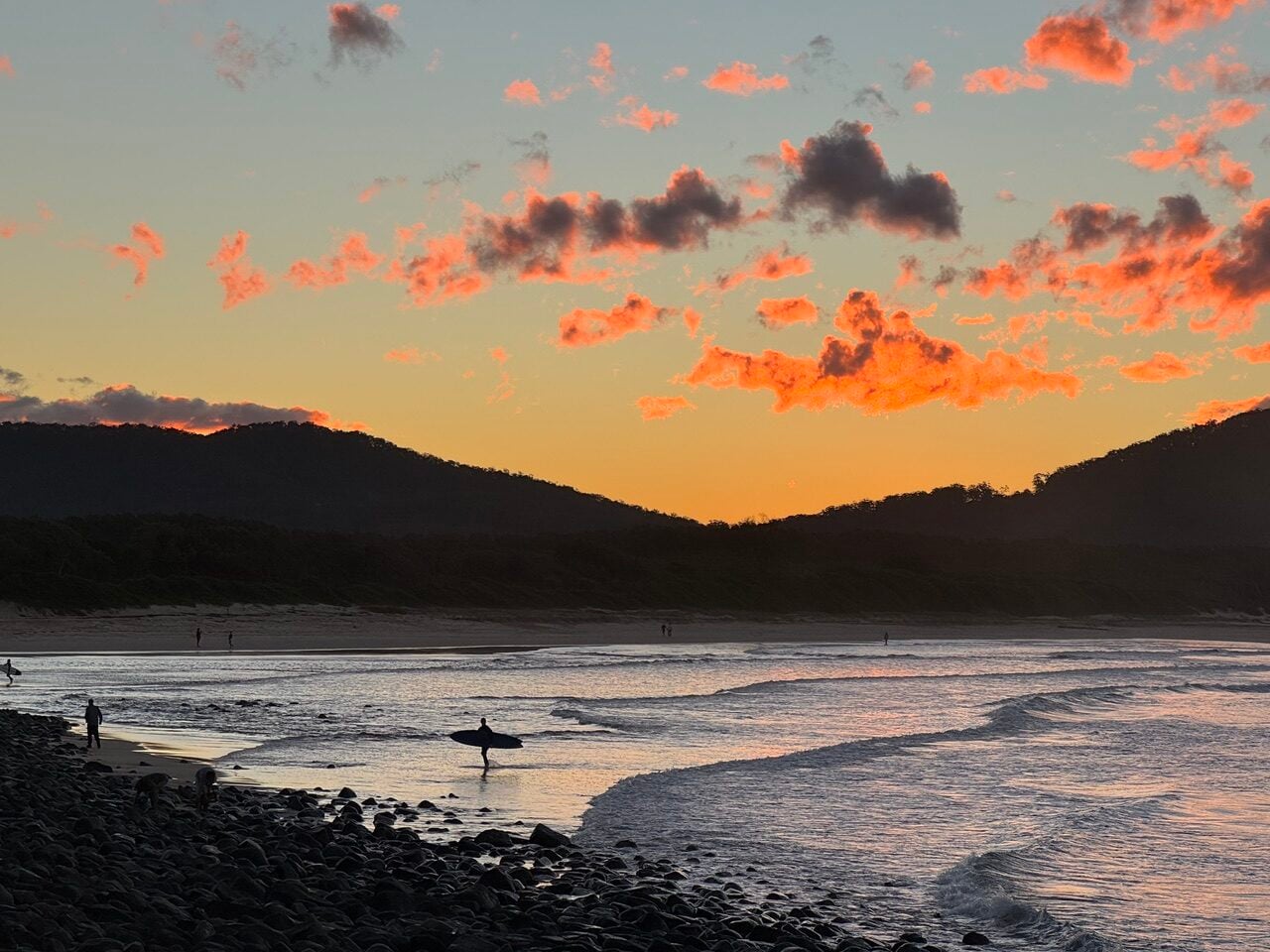 Silhouetted surfer walking along a rocky beach at sunset with orange clouds above rolling waves.