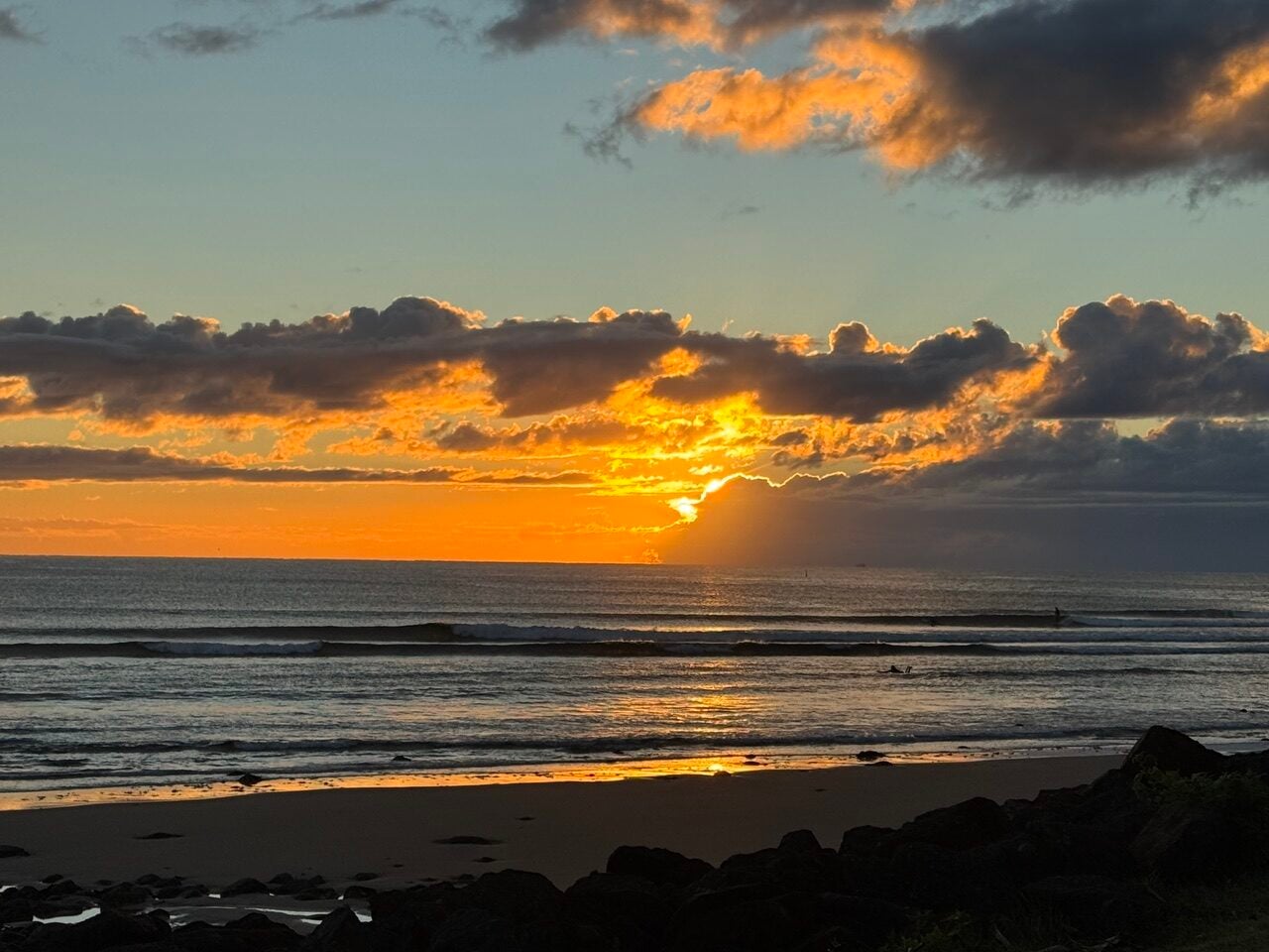 Sunset over the ocean with orange-cloud sky and gentle waves, rocky shore in the foreground.