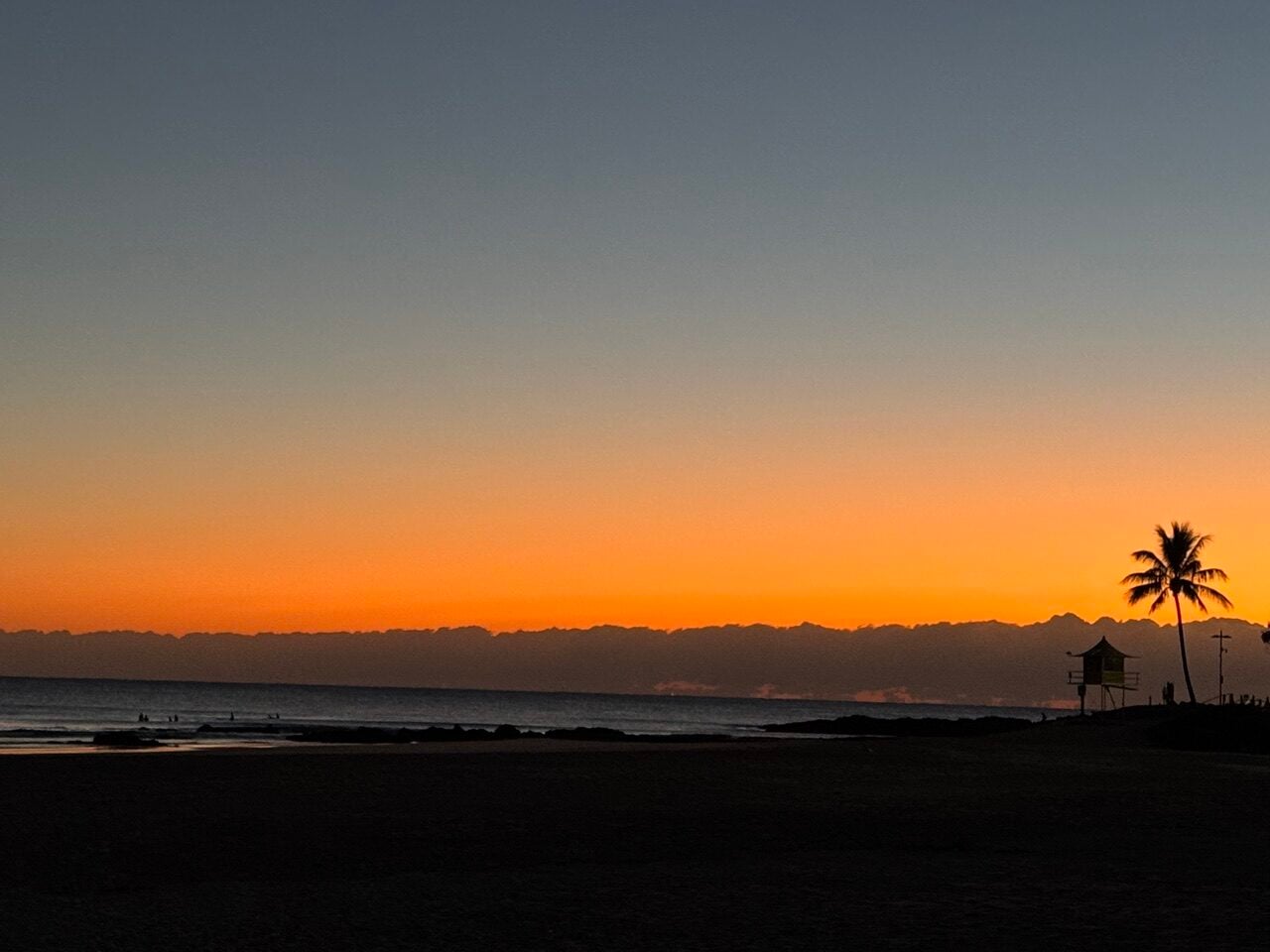 Sunset over the ocean with a palm tree and a lifeguard tower silhouetted on the right.