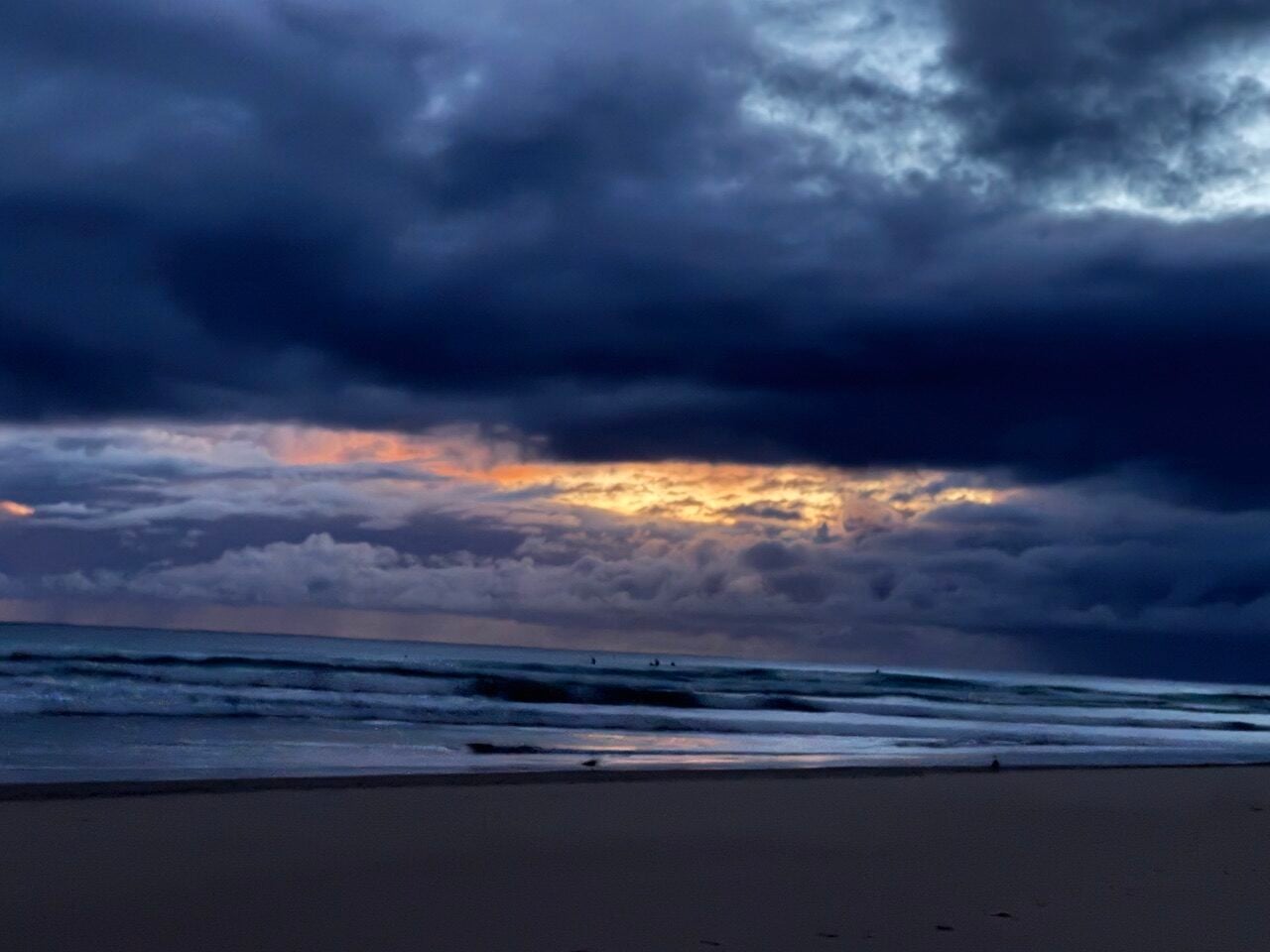 Dark storm clouds over a choppy sea at a deserted beach, with a narrow orange sunset glow on the horizon.