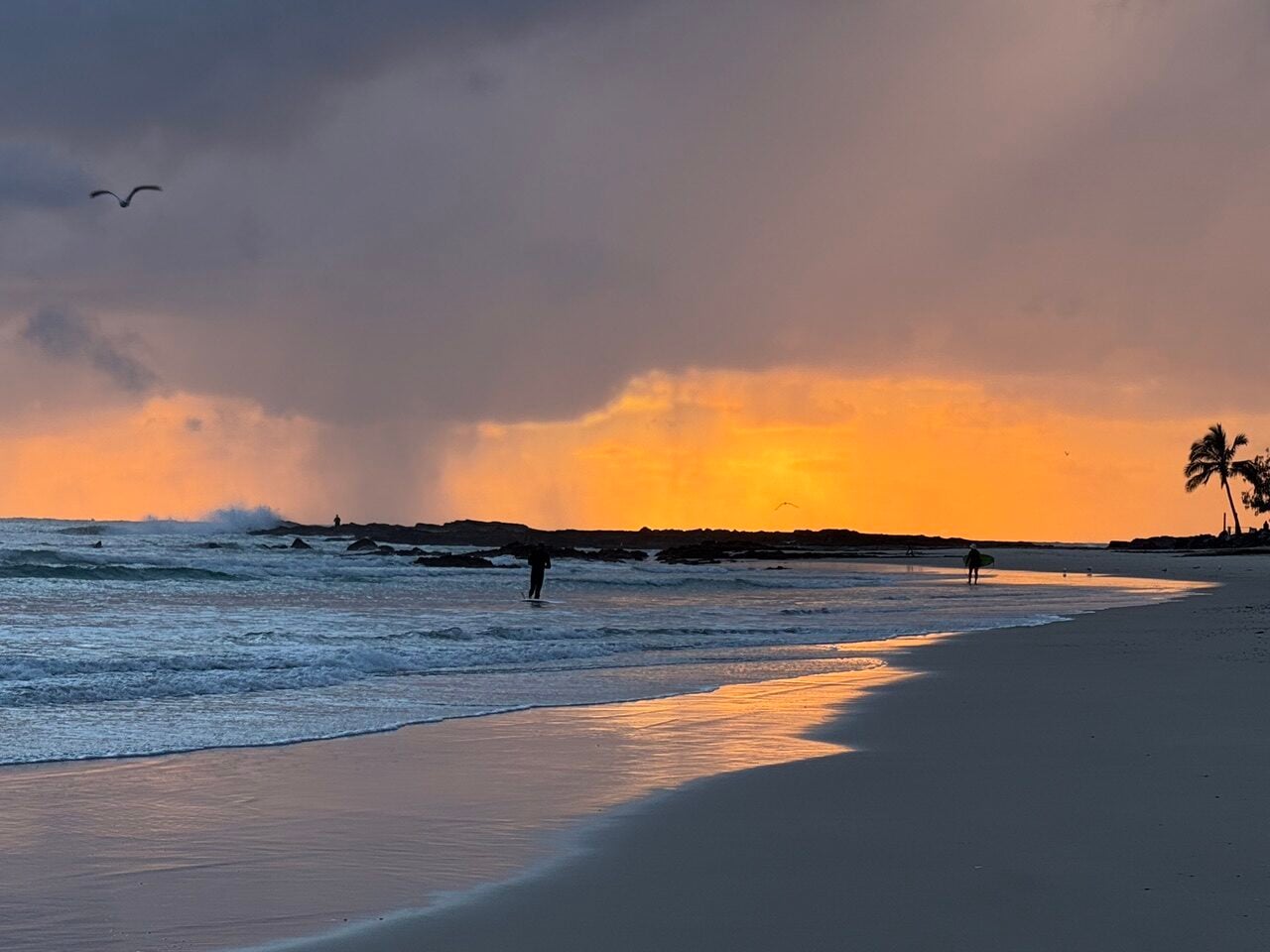 Sunset beach scene with orange sky, gentle waves, and two surfers walking along the shore near rocks.
