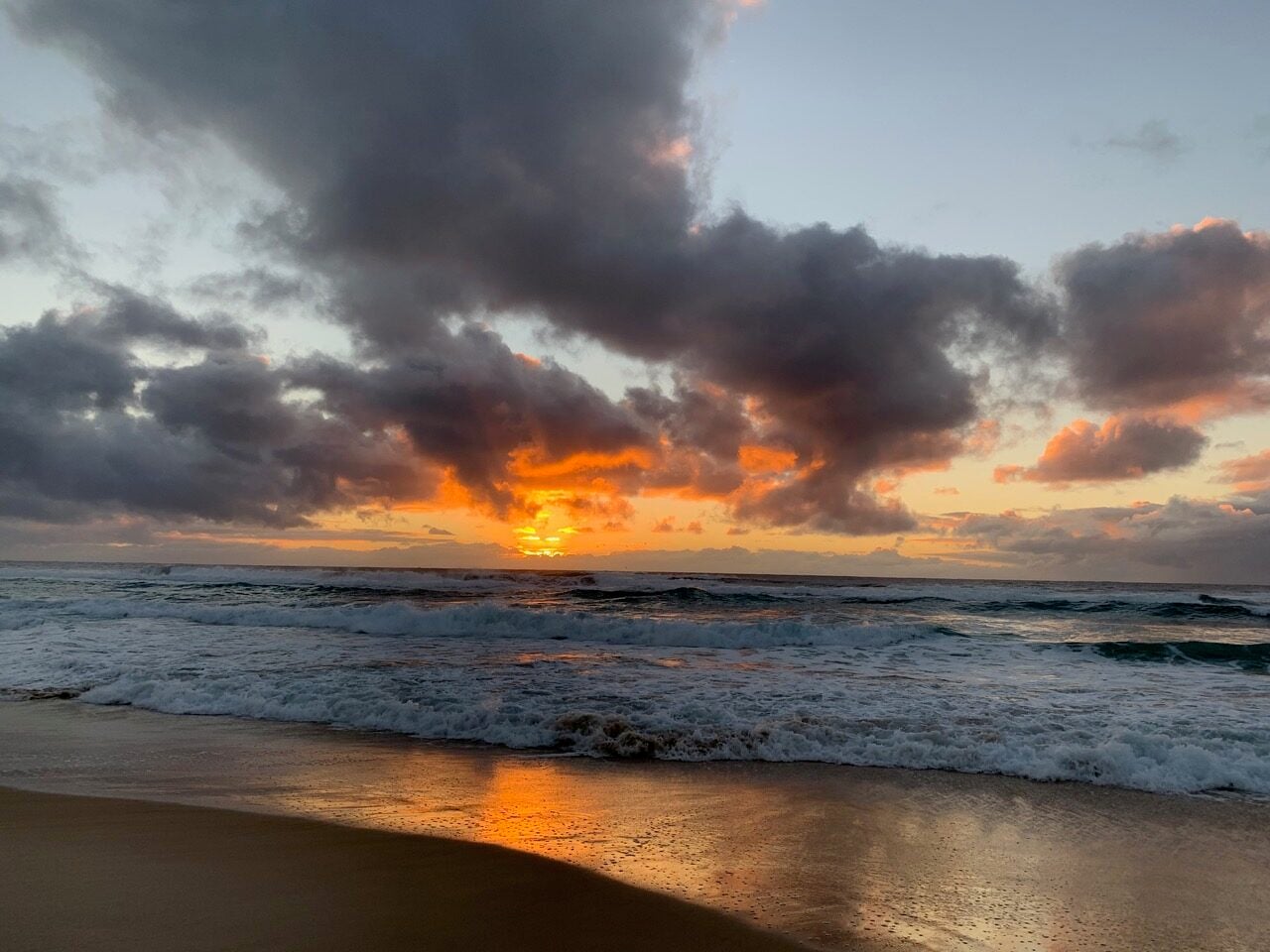 Sunset over the ocean with orange glow near the horizon and waves rolling onto a sandy beach.