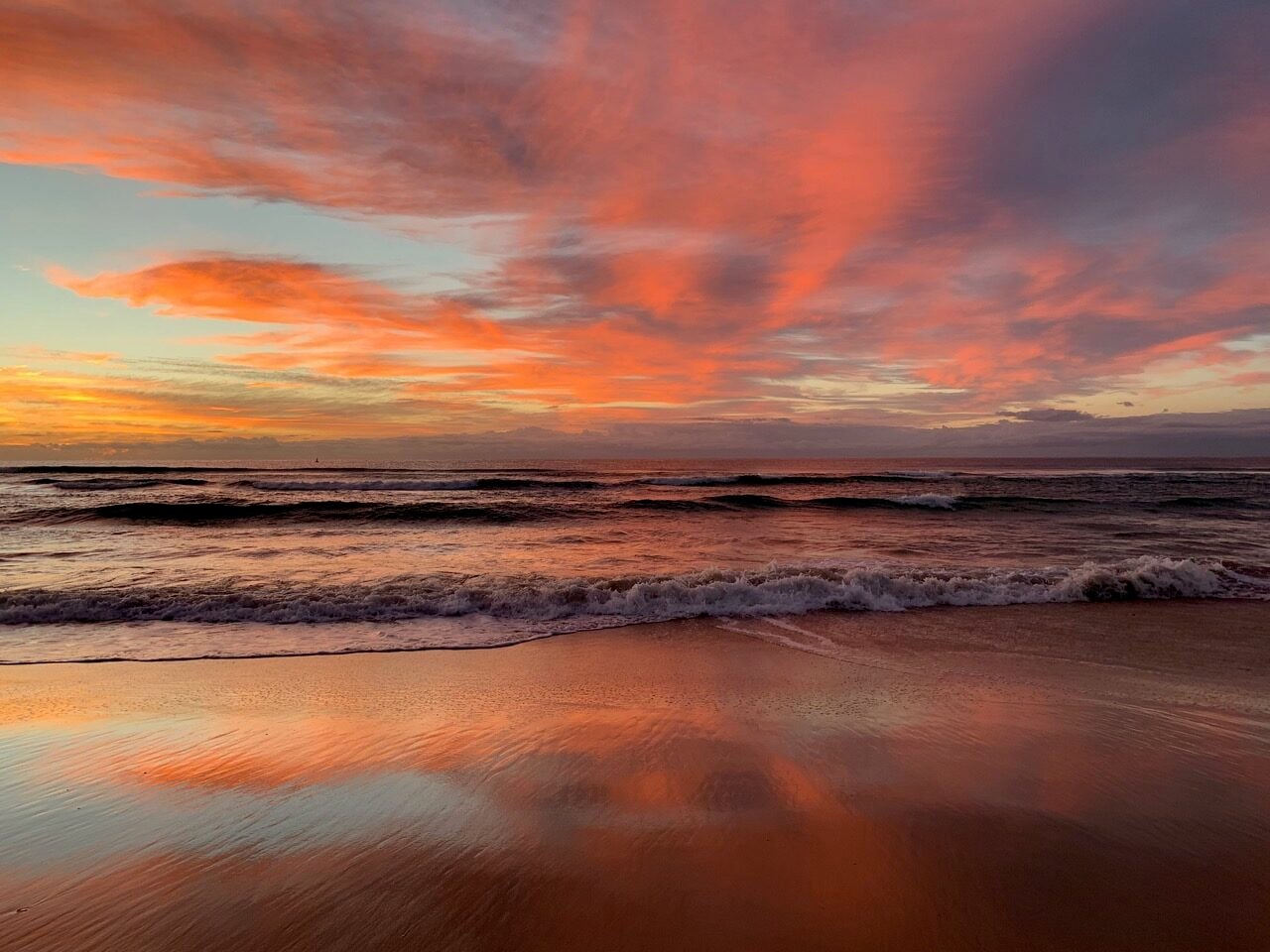 Sunset over the ocean with pink-orange clouds and waves washing onto a wet, reflective shoreline.