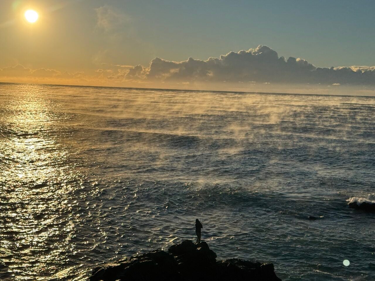 Silhouette of a person fishing on dark rocks by the ocean at sunset, with golden reflections on the water and a bright sun low in the sky.