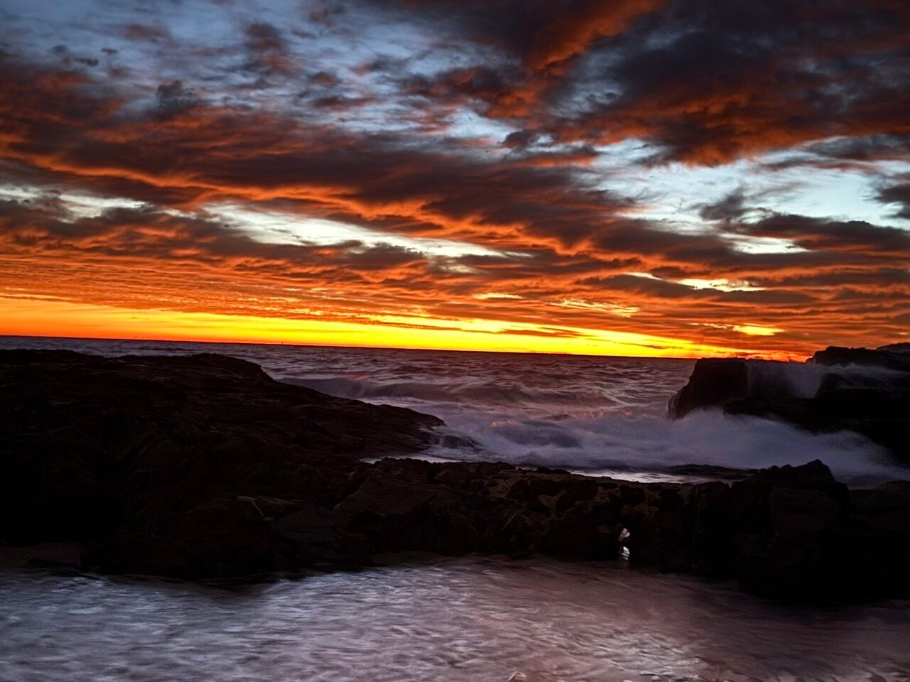 Sunset over a rocky shoreline with orange and red clouds, waves crashing against dark rocks.