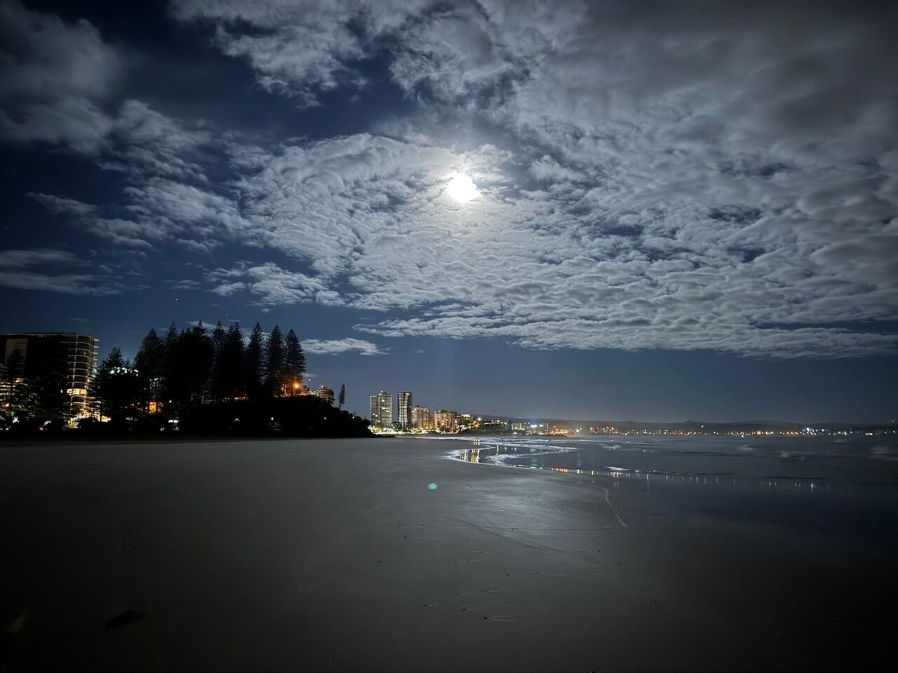 Moonlit night over a coastal city with illuminated buildings along the shore and a reflective, calm sea.