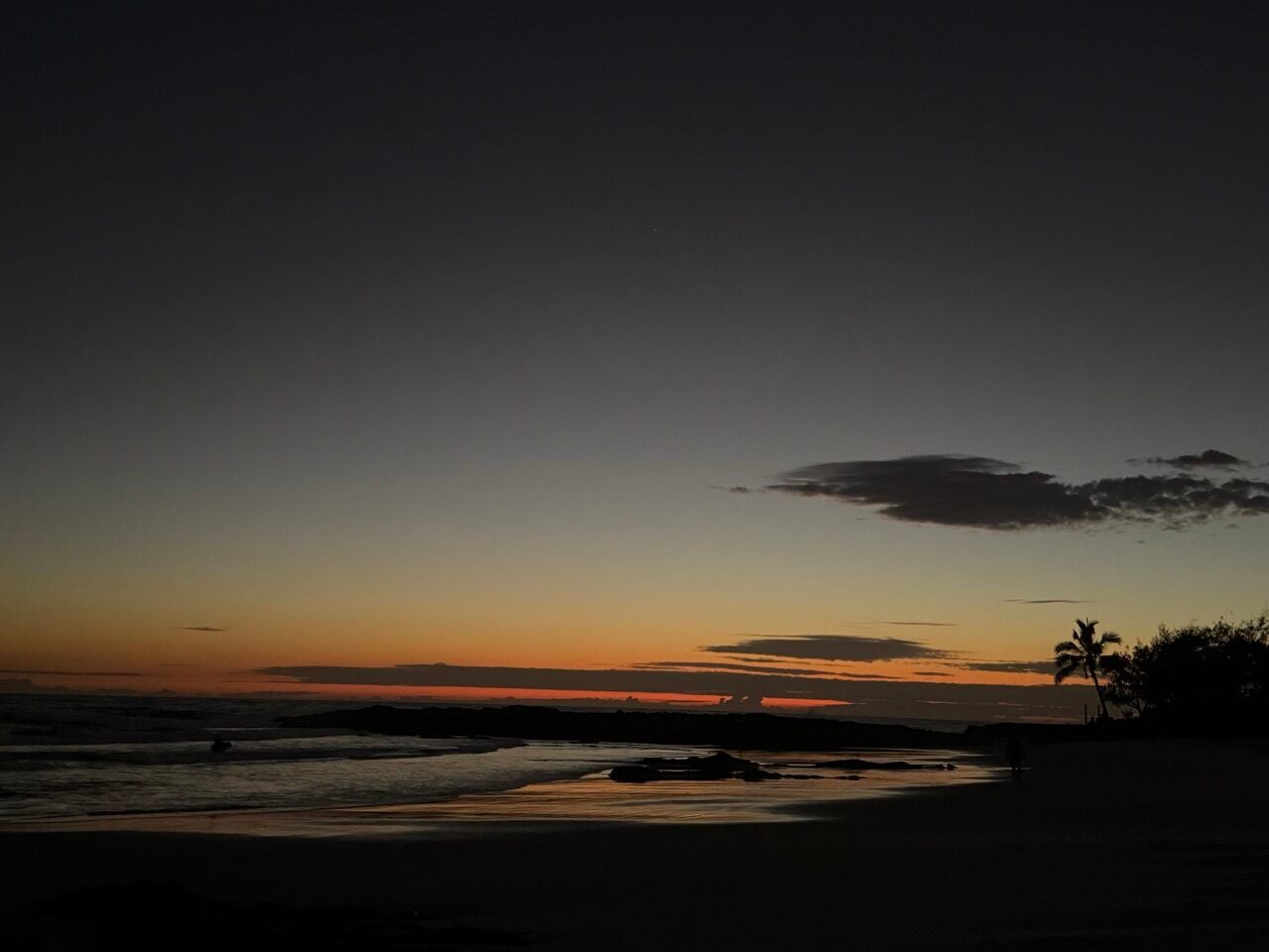 Silhouette of a tropical beach at sunset with a palm tree on the right, calm waves and an orange horizon.