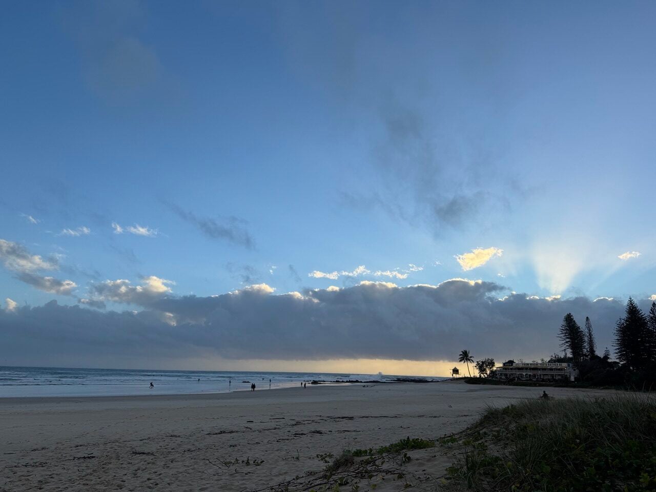 Wide beach scene at dusk with sunrays through clouds, calm ocean, and people strolling along the shoreline; buildings and palm trees visible on the right.