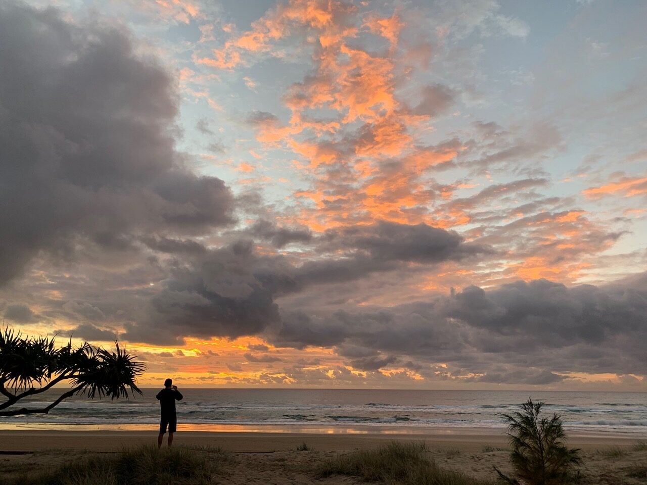 Silhouette of a person standing on a sandy beach at sunset, facing the ocean with dramatic orange clouds.