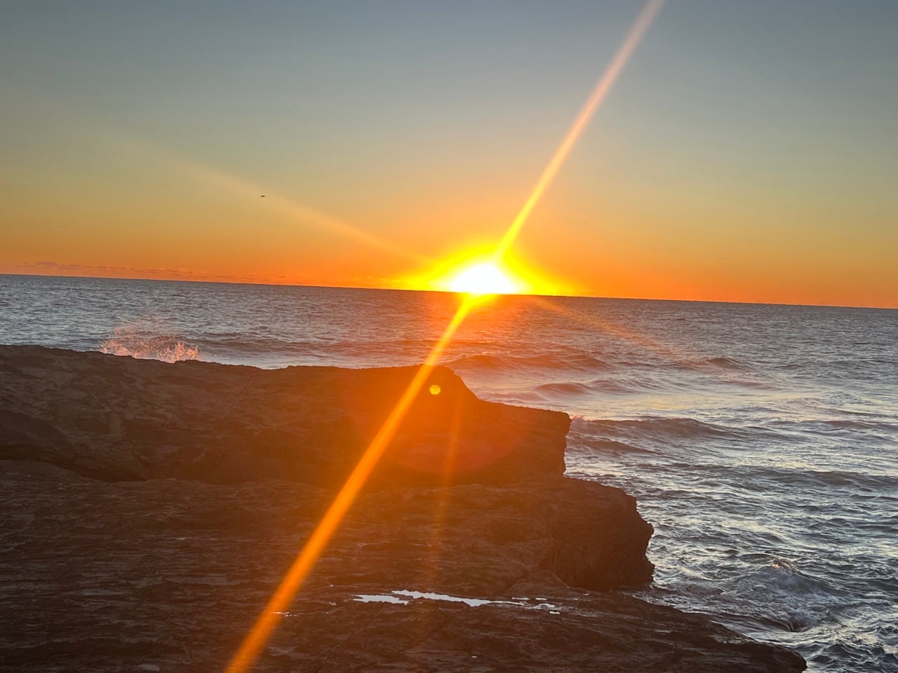 Sunset over the ocean behind a rocky coastline, with bright sun rays cutting across the scene.